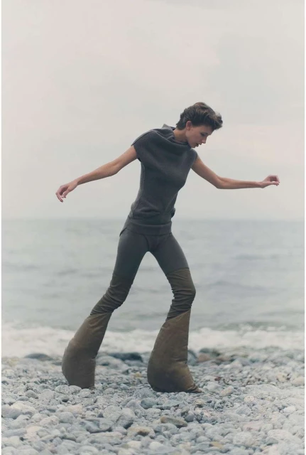 Woman on pebble beach wearing a gray outfit and large, padded shoes, walking by a gray ocean and overcast sky in an outdoor setting.