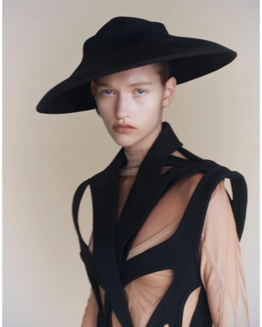 Headshot of a woman with pale skin wearing a black hat and top with structural cut-out detailing, set against a beige background.