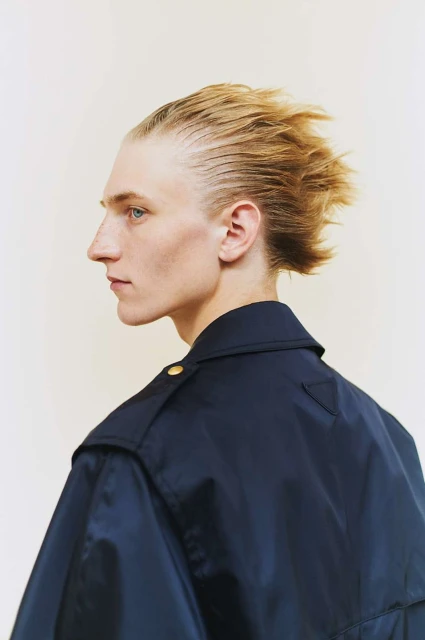 Profile of a blonde male model with styled hair wearing a navy blue jacket against an off-white backdrop in a studio setting.