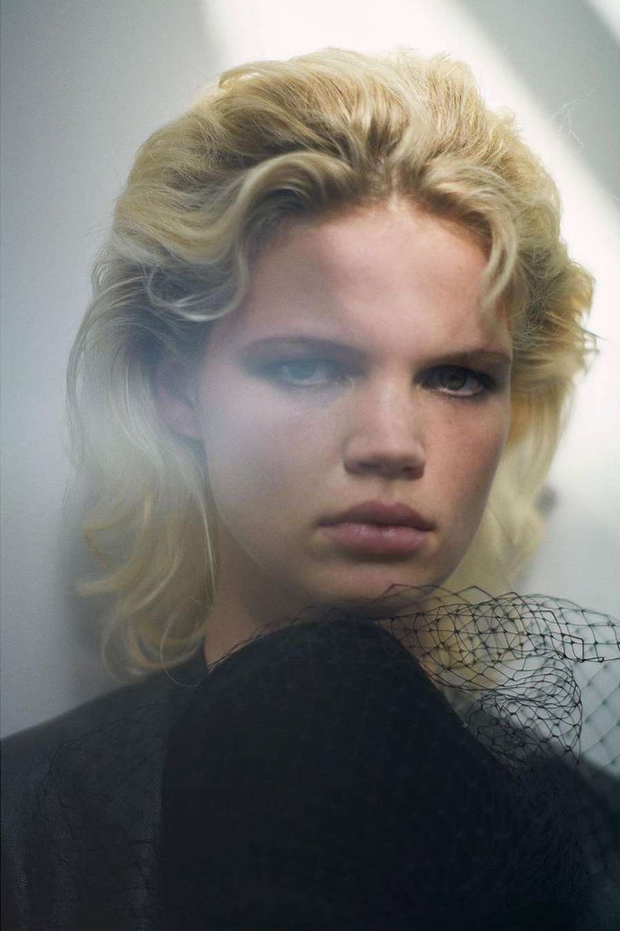 Close-up studio portrait of a woman with curly blonde hair, wearing a black top with a decorative black net veil.