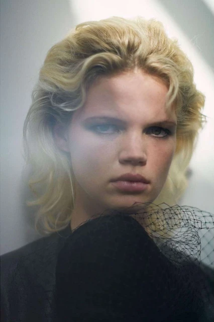 Close-up studio portrait of a woman with curly blonde hair, wearing a black top with a decorative black net veil.