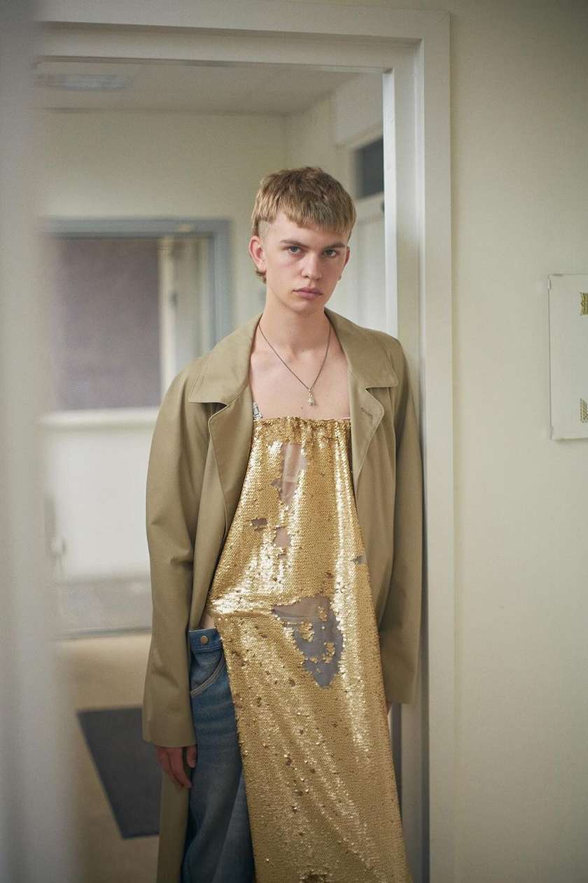 Young adult in a gold sequin dress and trench coat leans against a white door frame, posing in an indoor hallway with natural light.
