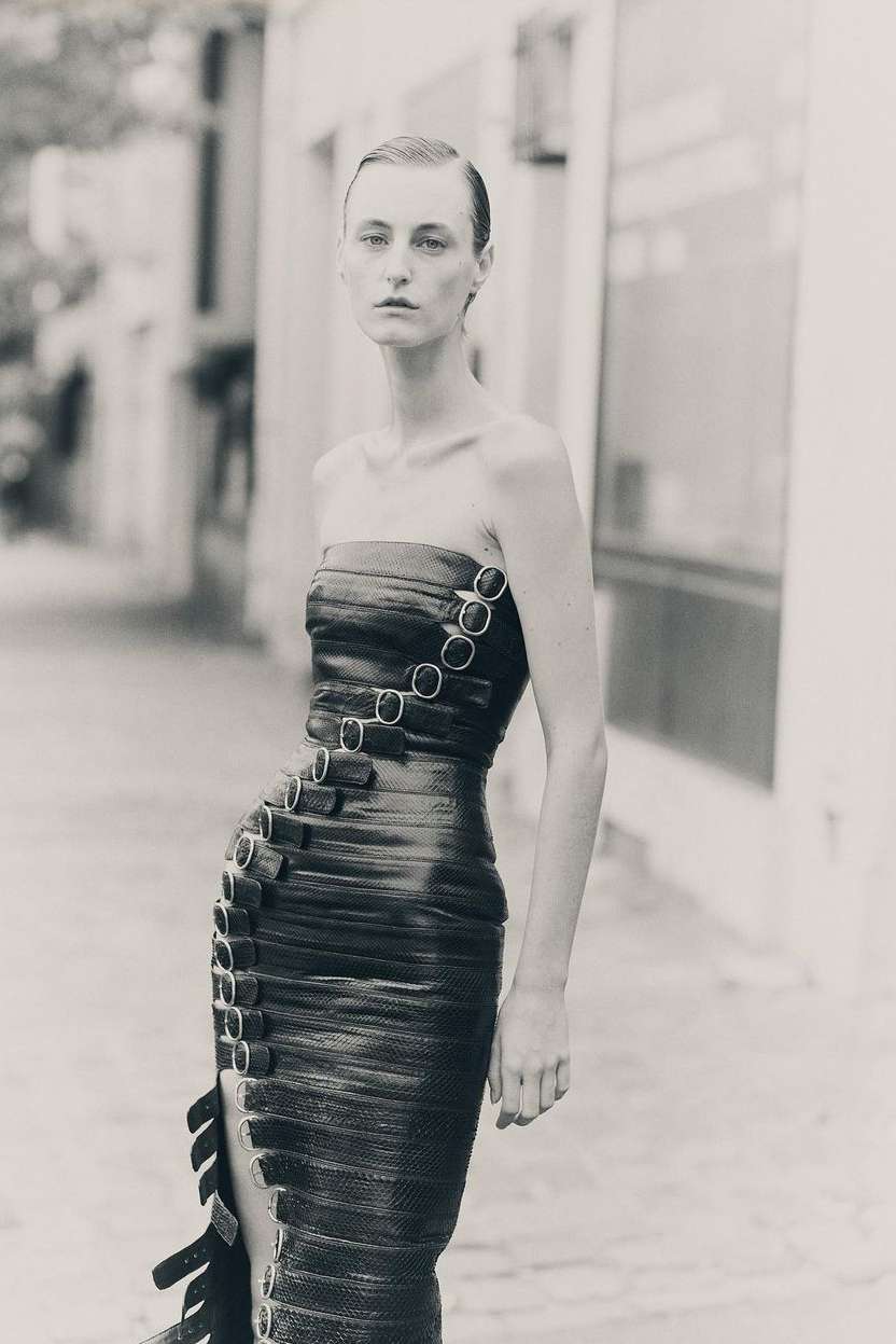 Adult female model in a structured, strapless dark dress with metal ring details, standing outdoors against an urban street background.