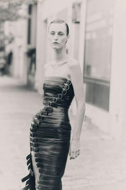Adult female model in a structured, strapless dark dress with metal ring details, standing outdoors against an urban street background.
