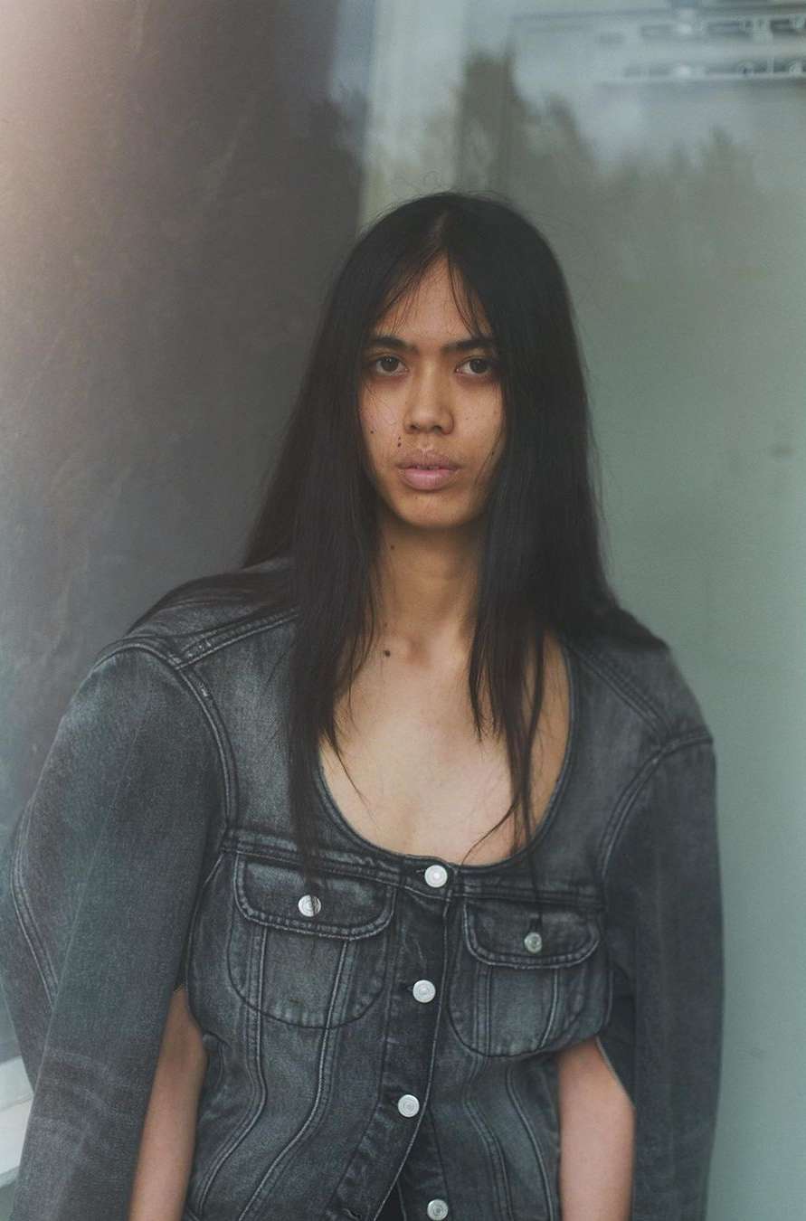 A stylish person with long dark hair wears a gray denim jacket against a blurred wall in a fashion-forward portrait.