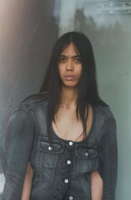 A stylish person with long dark hair wears a gray denim jacket against a blurred wall in a fashion-forward portrait.