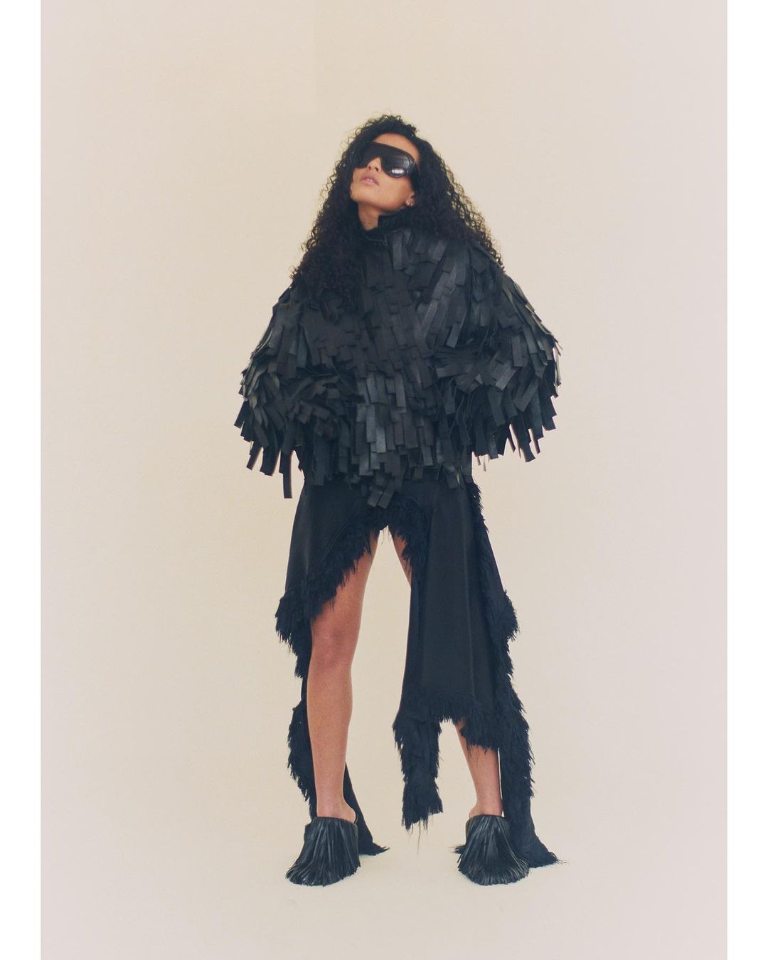 Woman with dark curly hair poses in a studio wearing a black fringed jacket, black skirt, sunglasses and tasseled heels in a studio.