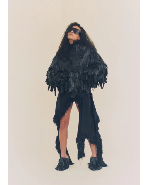 Woman with dark curly hair poses in a studio wearing a black fringed jacket, black skirt, sunglasses and tasseled heels in a studio.