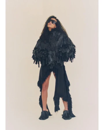 Woman with dark curly hair poses in a studio wearing a black fringed jacket, black skirt, sunglasses and tasseled heels in a studio.