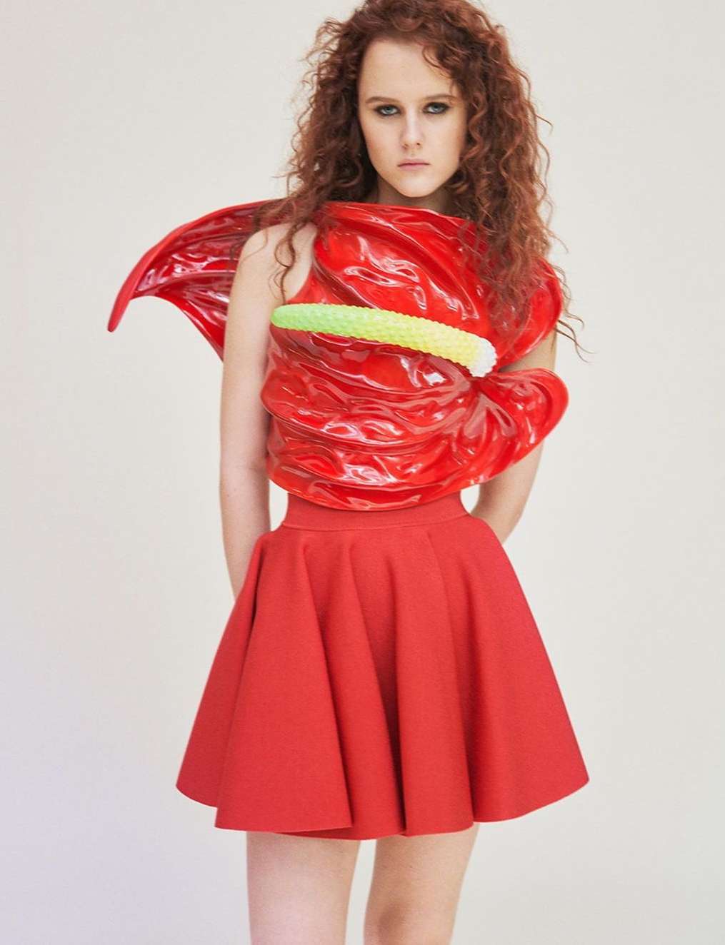 Woman in a red skirt and a top made from a red anthurium flower, posing in a studio with a neutral background.