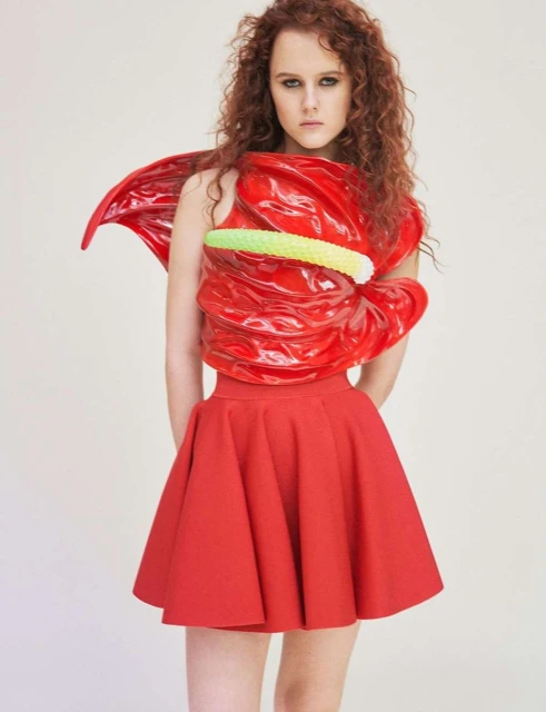 Woman in a red skirt and a top made from a red anthurium flower, posing in a studio with a neutral background.