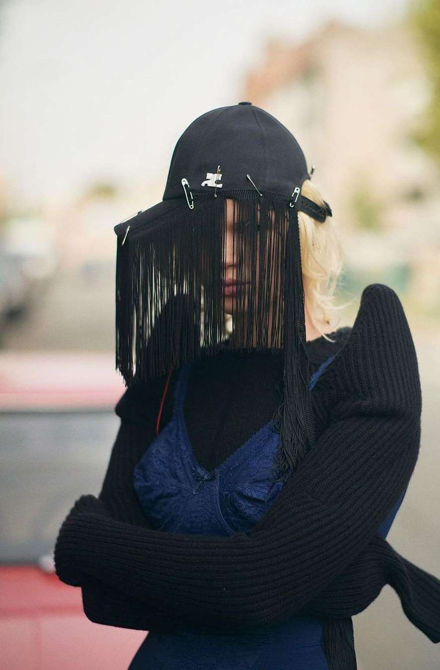 Fashionable woman wearing a black fringe baseball cap, dark clothing, and blue denim top posing in a blurred outdoor setting.