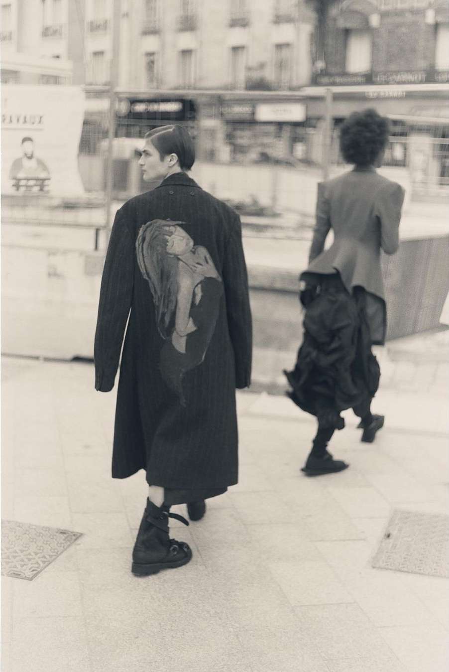 Two people walking on a city sidewalk, one wearing a long black coat with an illustration and the other a deconstructed skirt.