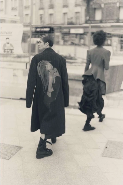 Two people walking on a city sidewalk, one wearing a long black coat with an illustration and the other a deconstructed skirt.