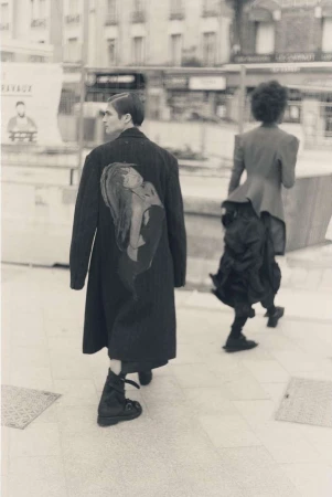 Two people walking on a city sidewalk, one wearing a long black coat with an illustration and the other a deconstructed skirt.