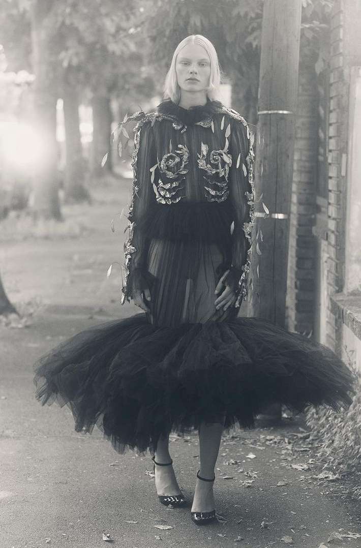 Woman in avant-garde black dress poses on sidewalk in black and white photo. Fashion editorial showcases unique style, tulle skirt, and heels.