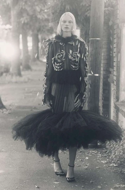 Woman in avant-garde black dress poses on sidewalk in black and white photo. Fashion editorial showcases unique style, tulle skirt, and heels.