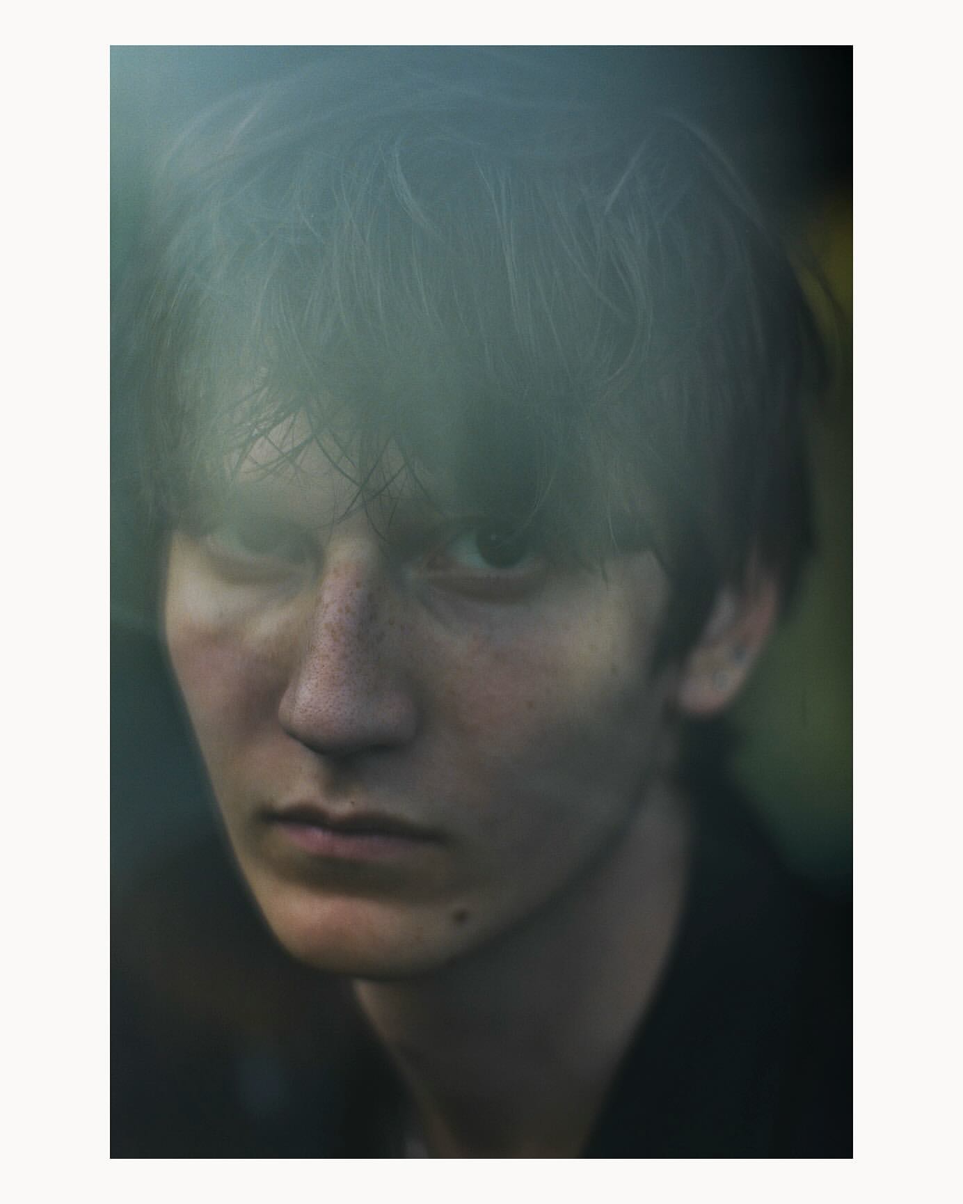 Close-up portrait of a young adult male with dark hair and a serious expression, set against a blurred, natural background with soft lighting.