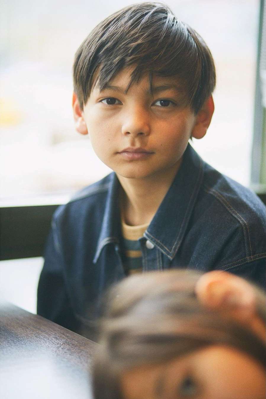 Portrait of a young boy wearing a dark denim jacket with another person visible in the foreground by photographer Ola Rindal.