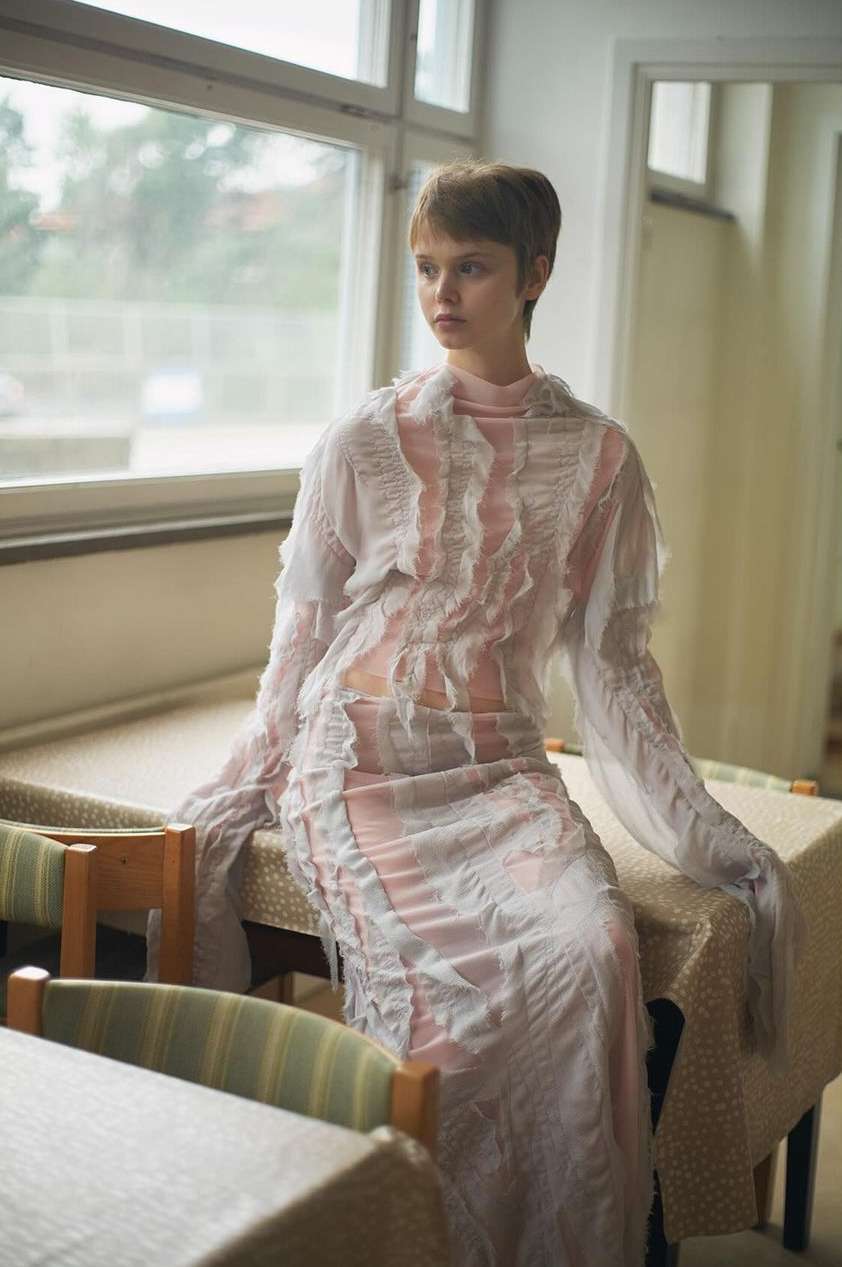 A woman with short brown hair wearing a sheer ruffled dress seated on a patterned table inside a room.