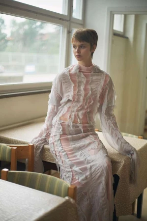 A woman with short brown hair wearing a sheer ruffled dress seated on a patterned table inside a room.