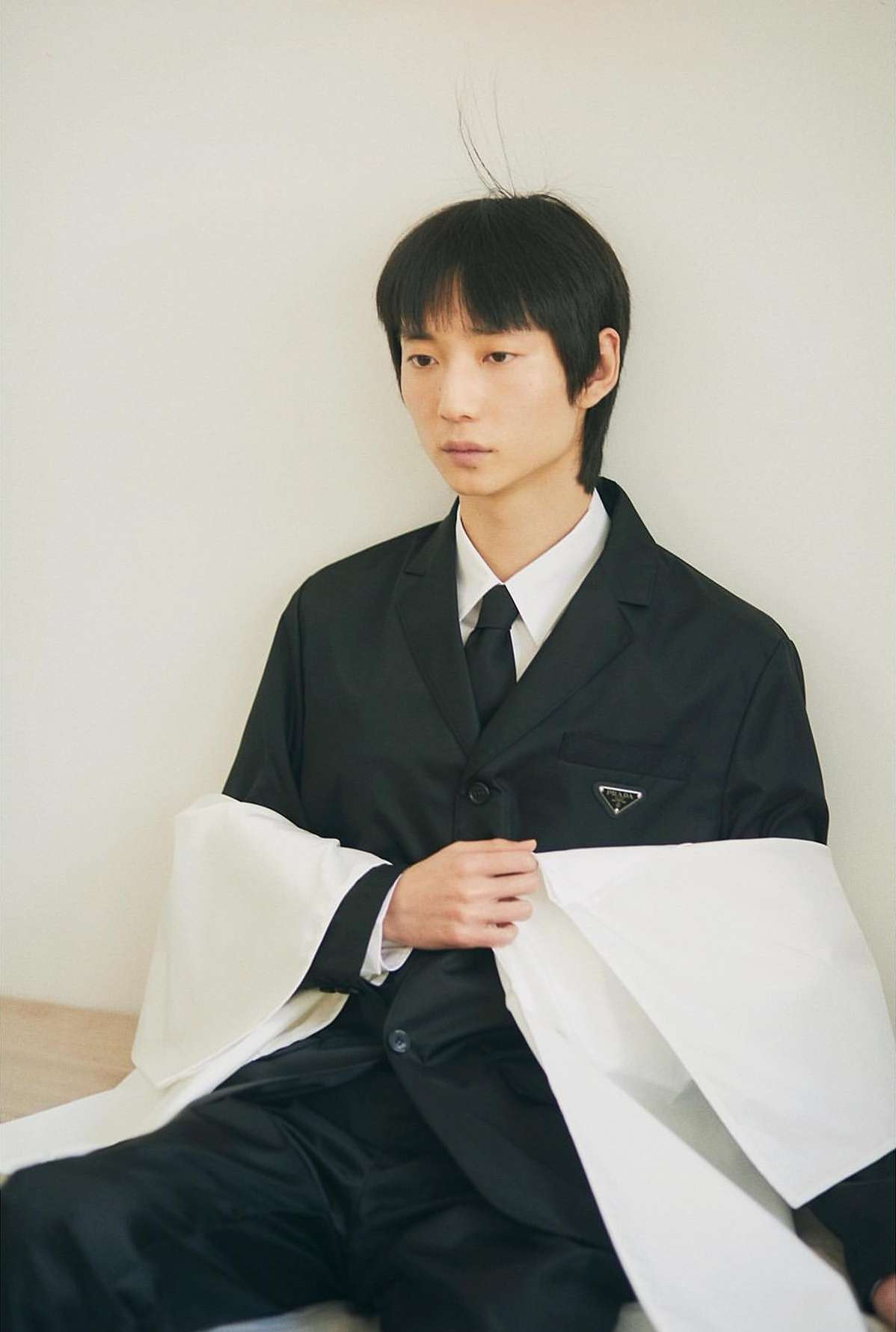 Young Asian male model poses indoors wearing black Prada suit with a white fabric drape, showcasing editorial fashion.
