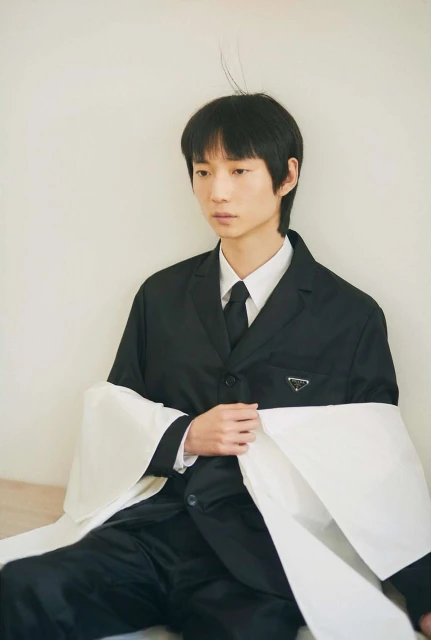 Young Asian male model poses indoors wearing black Prada suit with a white fabric drape, showcasing editorial fashion.
