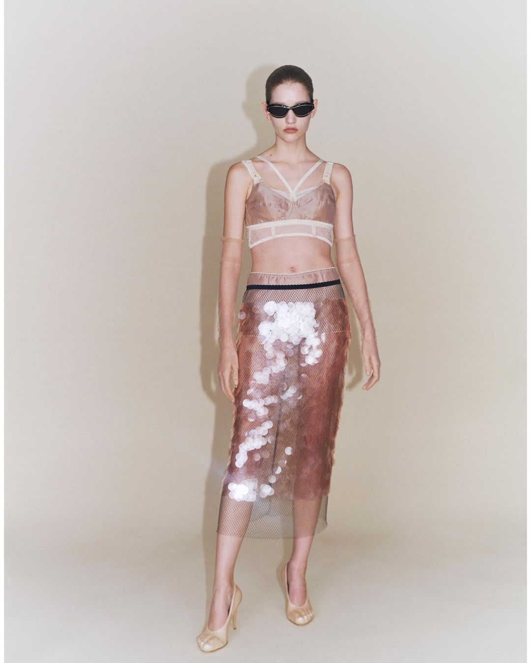 Full-length studio shot of a woman wearing a nude-tone bra top, semi-sheer skirt with metallic sequins, sunglasses, and nude high heels.