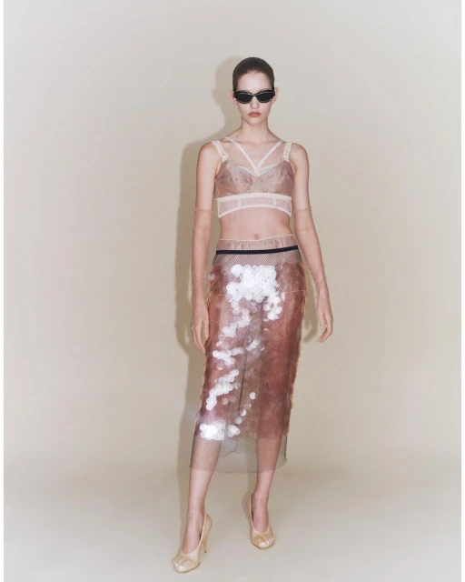 Full-length studio shot of a woman wearing a nude-tone bra top, semi-sheer skirt with metallic sequins, sunglasses, and nude high heels.