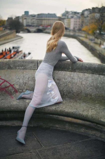Woman in sheer skirt poses on stone ledge overlooking the Seine in Paris, with neutral tones and city backdrop, fashion forward style.
