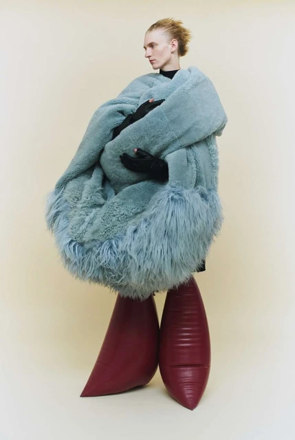 Androgynous model in a powder blue faux fur coat and textured burgundy platform shoes posing in a neutral colored studio.