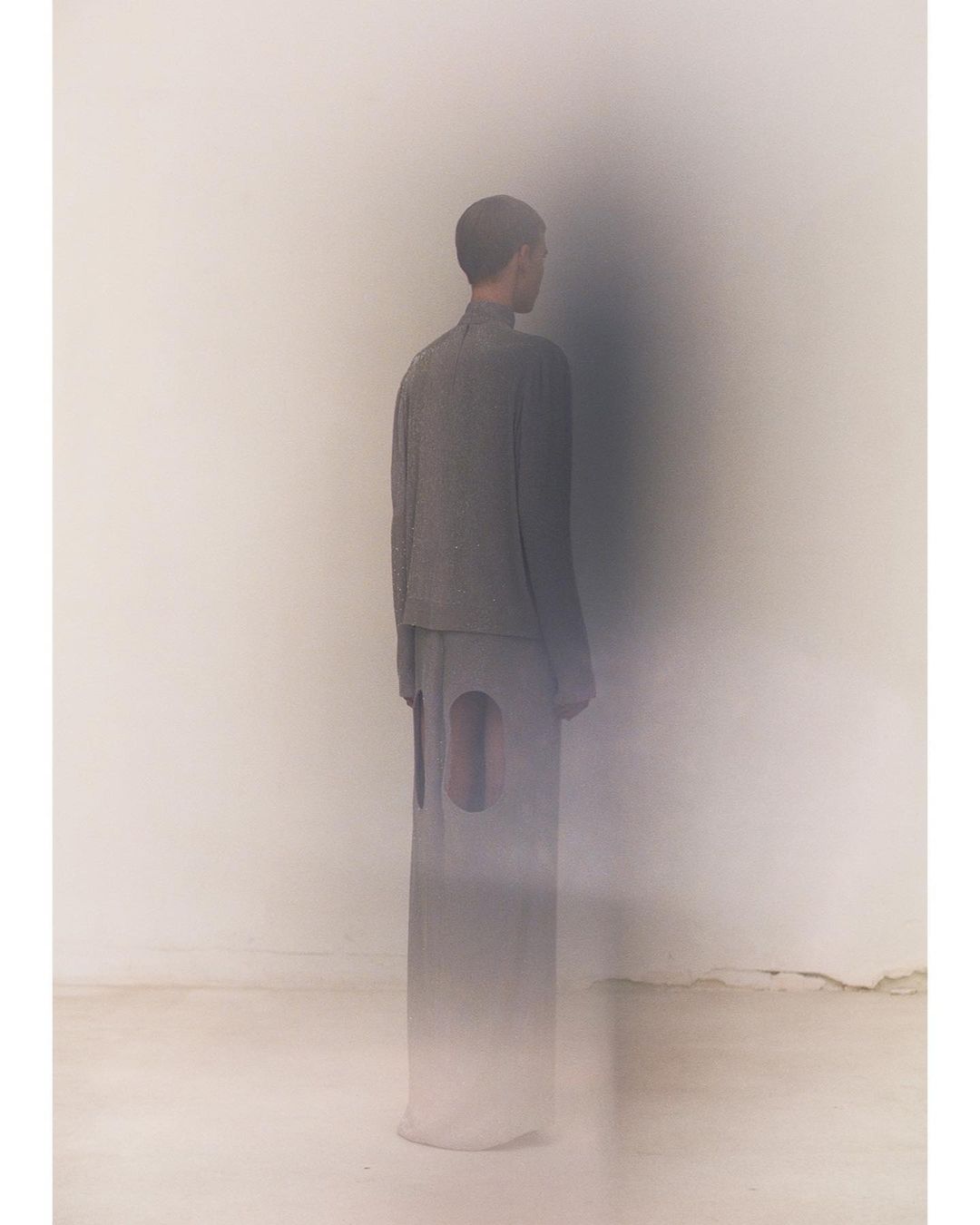 Fashion shot of a person in gray clothing standing in front of a white wall with a blurred shadow, minimalist style, studio lighting.