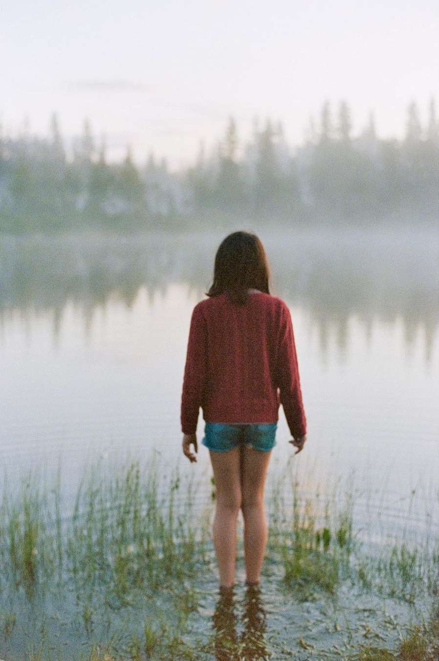 Young woman standing in shallow lake water facing away from the camera toward a misty tree line during dusk.