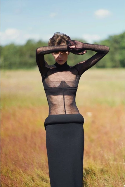 Woman in sheer top and long black skirt stands in field with hands over face, bathed in natural light with trees in background.