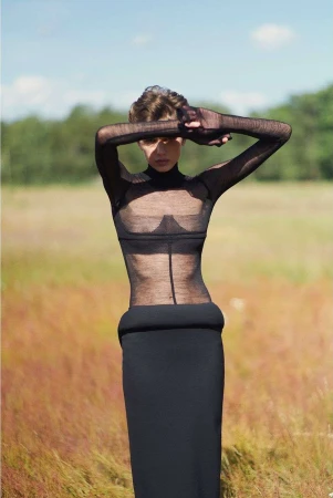 Woman in sheer top and long black skirt stands in field with hands over face, bathed in natural light with trees in background.