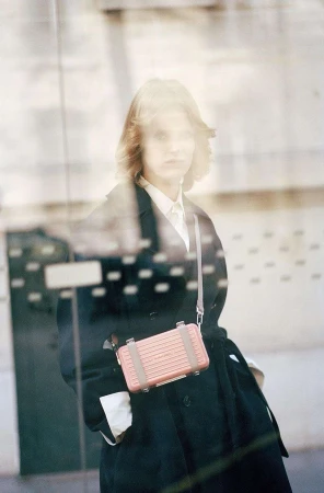 Woman wearing a black coat and white shirt, carrying a pink crossbody bag, viewed through a glass window in Paris by Ola Rindal.