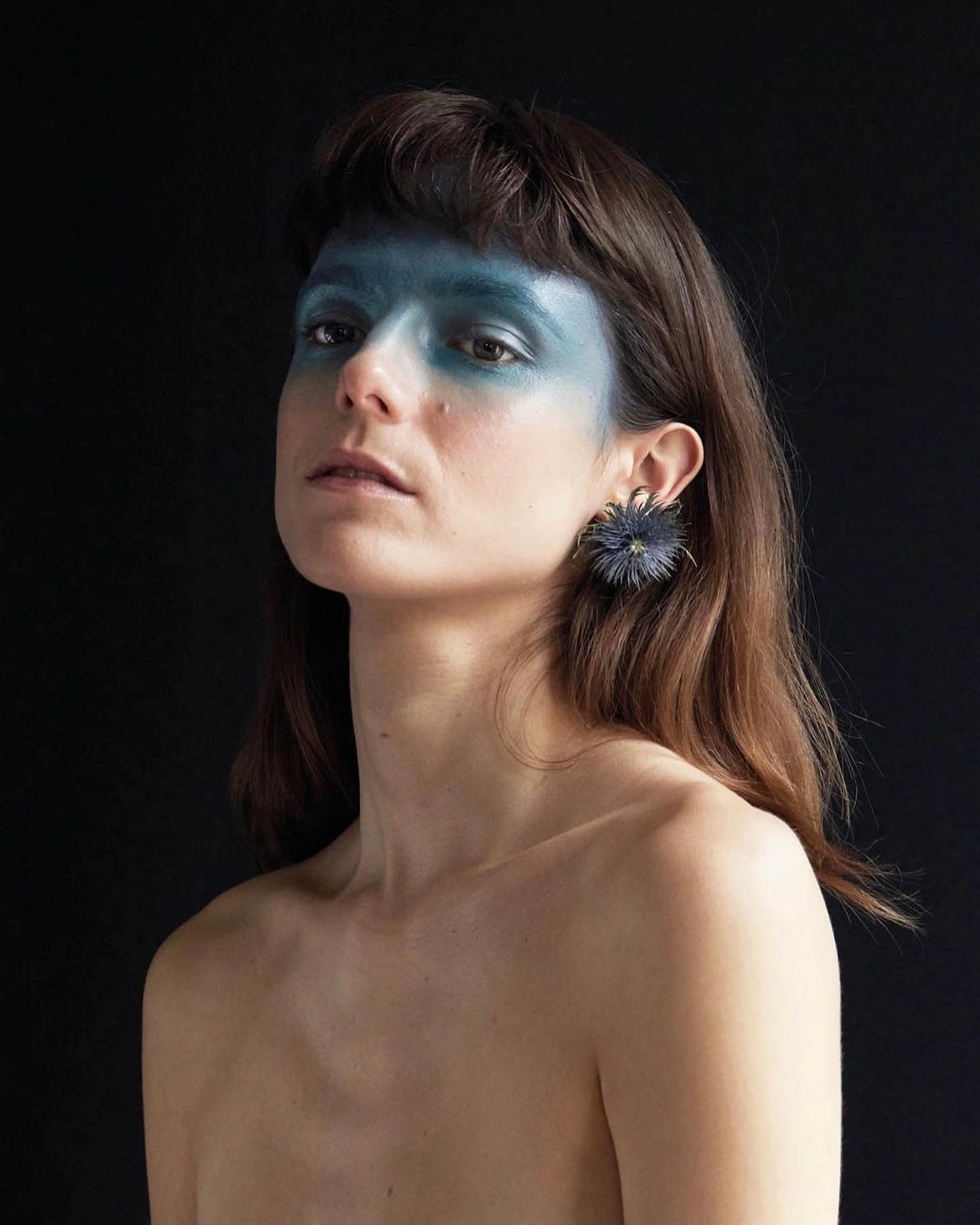 Portrait of a woman with blue eye makeup and bare shoulders against a black background, posing with an earring.