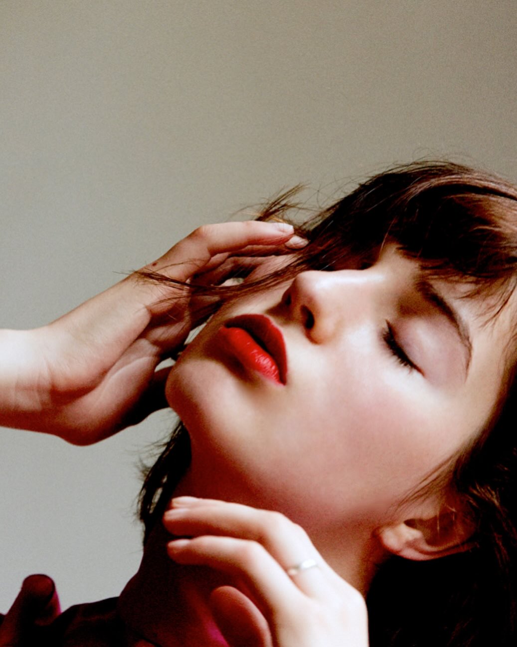 Portrait of an adult woman with red lipstick and closed eyes, hands near her face in soft lighting, against a neutral backdrop.