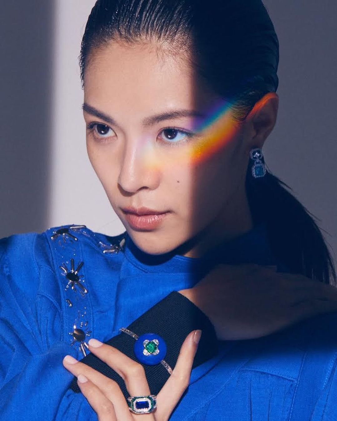 Asian woman with jewelry is captured in a medium shot, showcasing a bright blue blouse and rainbow lighting across her face in a studio.