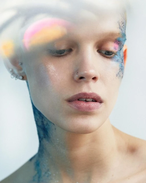 Close-up portrait of a young adult female with blue glitter makeup on her face and neck against a blurred light background.
