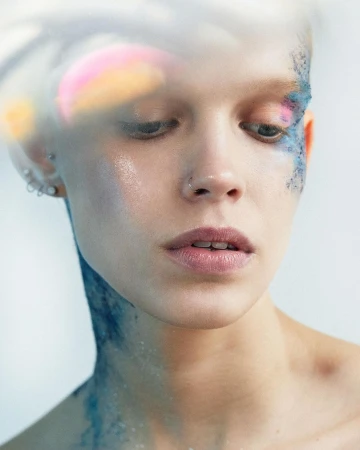 Close-up portrait of a young adult female with blue glitter makeup on her face and neck against a blurred light background.