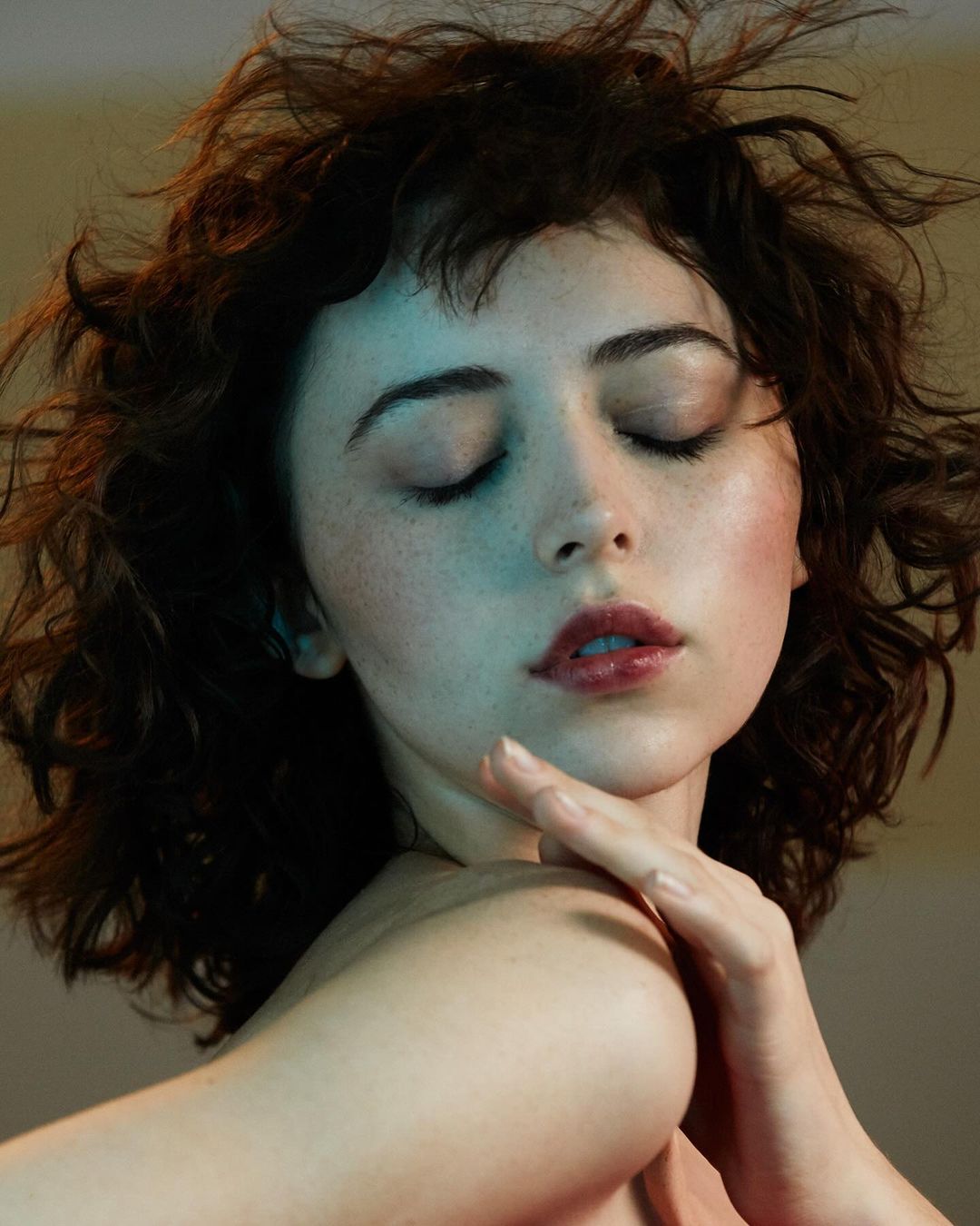 Portrait of an adult woman with closed eyes, brown curly hair, and soft lighting accentuating facial features and skin texture.