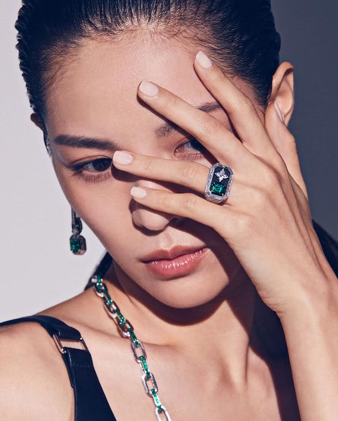 Close-up portrait of an Asian woman with dark hair and fair skin, wearing an ornate ring and earrings with green stones.