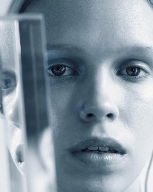 Close-up portrait of a woman with fair skin, looking directly at the camera with a serious expression, framed by a reflective surface.