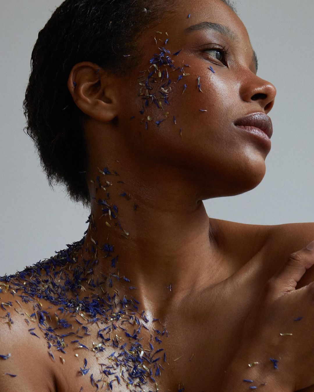 Close-up studio shot of an adult female model with a bare chest and blue flower petals on her face, chest, and shoulders.