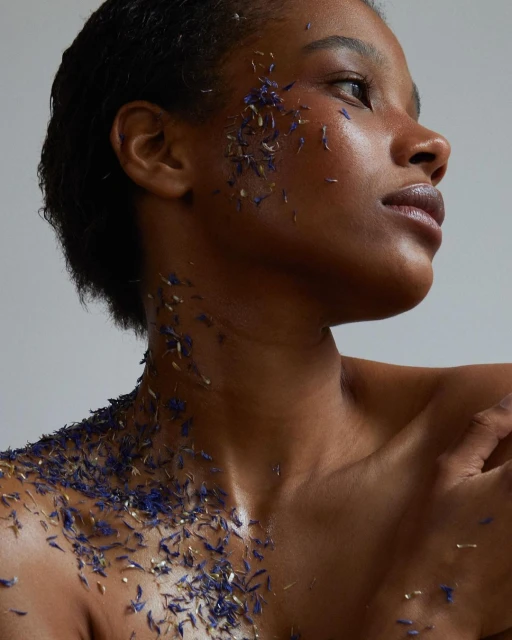 Close-up studio shot of an adult female model with a bare chest and blue flower petals on her face, chest, and shoulders.