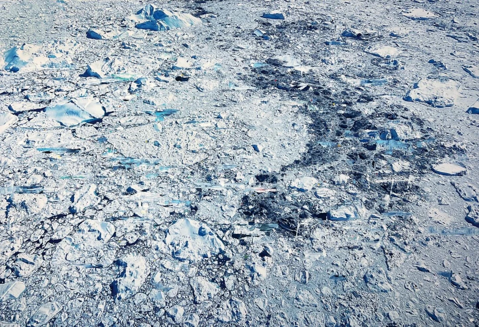 Aerial view of a vast, icy landscape featuring ice floes and dark patches, creating a textured, natural pattern in a cold environment.