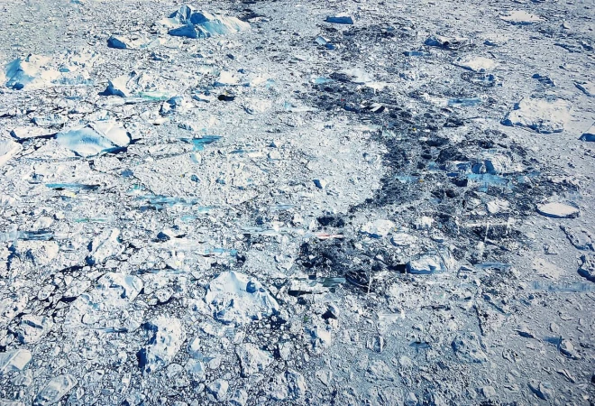 Aerial view of a vast, icy landscape featuring ice floes and dark patches, creating a textured, natural pattern in a cold environment.