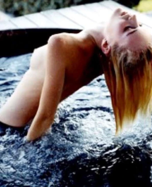 Blonde woman with wet hair leans back into the swirling water of a tub in a close-up portrait, outdoor, showing bare shoulders.