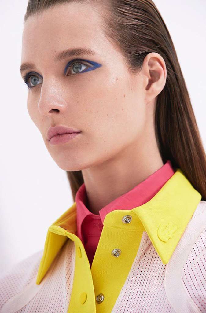 Close-up shot of a woman with blue eyeshadow wearing a yellow and pink collared shirt on a white background in a beauty context.